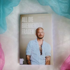 Das Buch "All die brennenden Fragen" liegt auf einem Hintergrund in den trans Pride Farben.