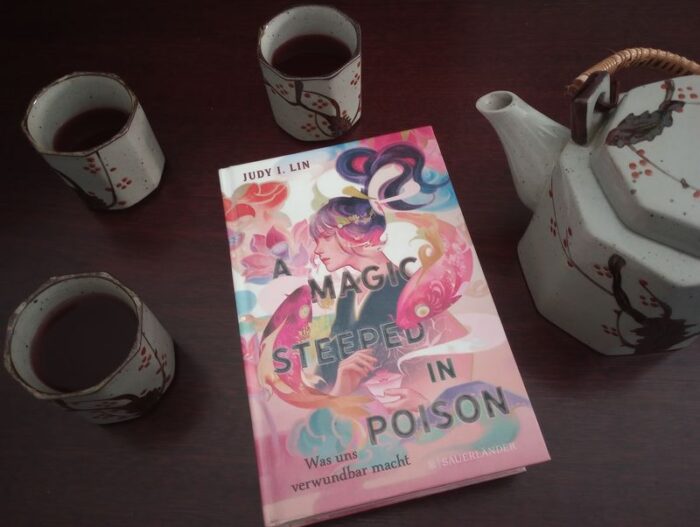 Das Buch "A magic steeped in poison" liegt auf einem dunklen Untergrund. Daneben stehen drei volle Teetassen und eine Teekanne