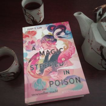 Das Buch "A magic steeped in poison" liegt auf einem dunklen Untergrund, daneben stehen eine Teekanne und Teetassen.