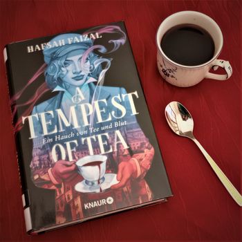 Das Buch "A tempest of tea" von Hafsah Faizal liegt auf einem dunkelroten Untergrund, daneben sind ein Teelöffel und eine Tasse mit dunkelroter Flüssigkeit.