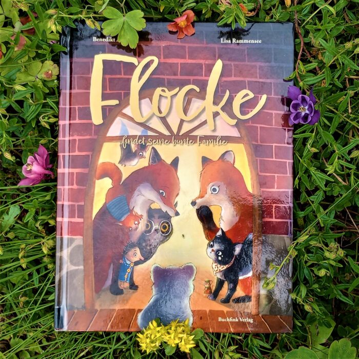 Ein Foto des Buchs "Flocke findet seine bunte Familie" in einem Blumenbeet. Über das Foto führt ein Link zum Buch im Katalog der Stadtbücherei.