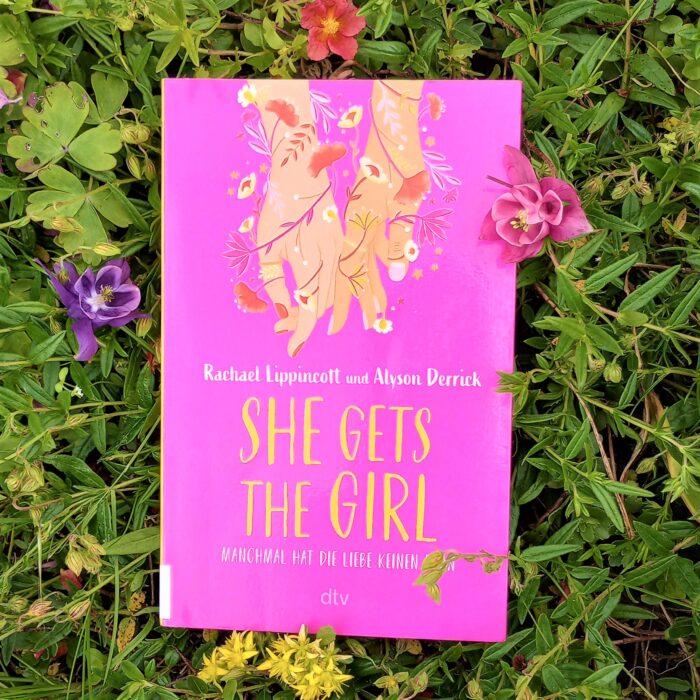 Ein Foto des Buchs "She gets the girl" in einem Blumenbeet. Über das Foto führt ein Link zum Buch im Katalog der Stadtbücherei.