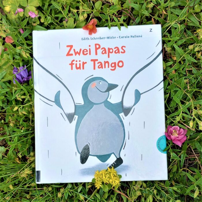 Ein Foto des Buchs "Zwei Papas für Tango" in einem Blumenbeet. Über das Foto führt ein Link zum Buch im Katalog der Stadtbücherei.