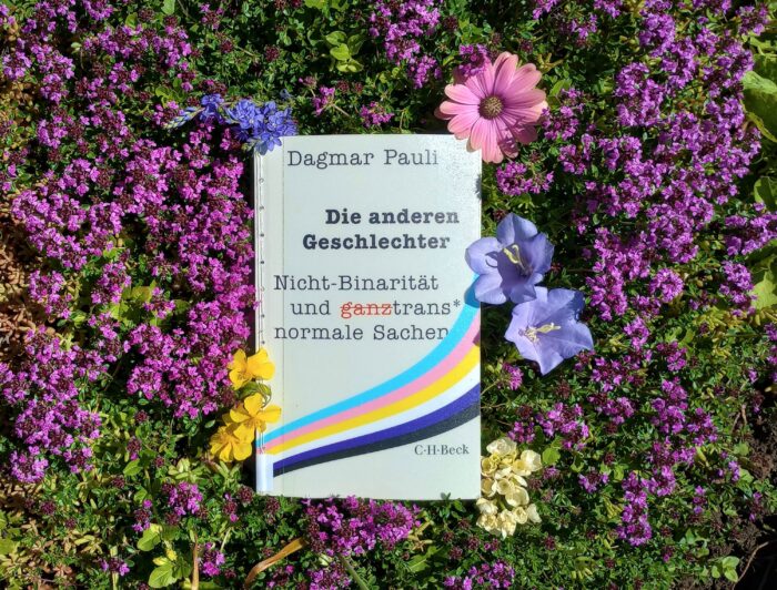 Das Buch "Die anderen Geschlechter" von Dagmar Pauli liegt auf einem Blumenbeet, umgeben von Blüten in den trans und nonbinären Pride-Farben weiß, lila, rosa, gelb und hellblau.