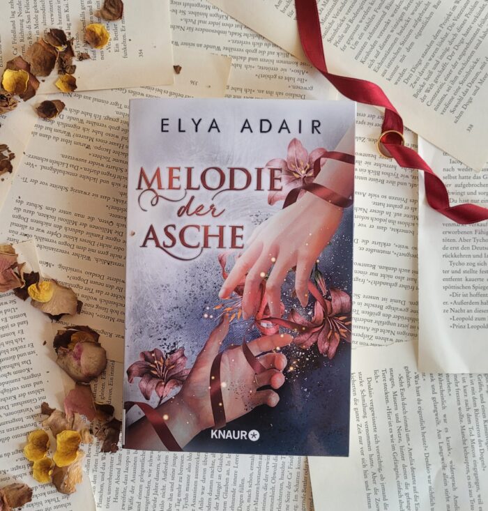 Das Buch Melodie der Asche liegt auf einem Hintergrund aus Buchseiten, Blumen und einem roten Band.