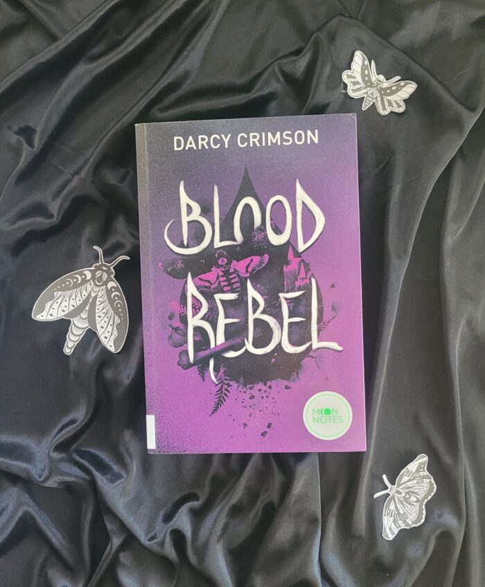 Das Buch "Blood Rebel" liegt auf einem Hintergrund aus schwarzem Samt, umgeben von Motten.