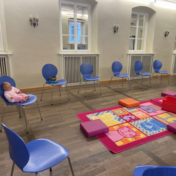 Auf dem Bild sieht man einen Stuhlkreis. In der Mitte befindet sich ein großer Teppich mit Sitzkissen, auf welchem die Kinder spielen können.