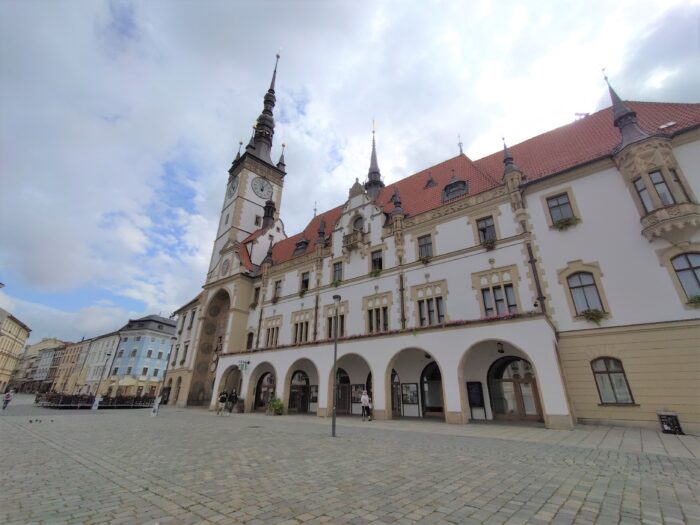 Ein Bild eines historischen Gebäudes in Olomouc. Es hat Bogengänge und einen Turm mit einer Turmuhr.