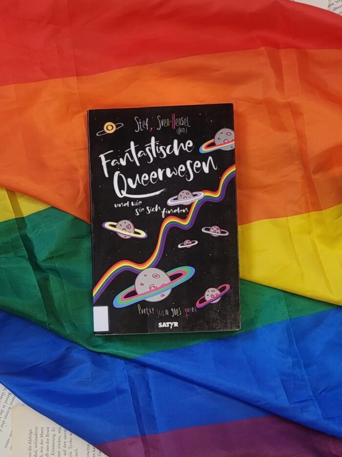 Das Buch "Fantastische Queerwesen und wie sie sich finden" liegt auf einer Pride Flagge.