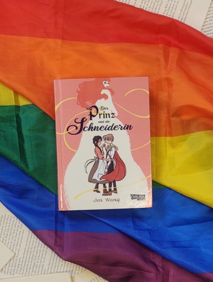 Das Buch "Der Prinz und die Schneiderin" liegt auf einer Pride Flagge.