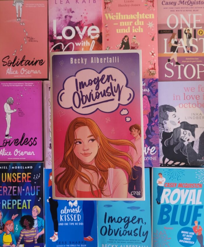Das Buch "Imogen, Obviously" liegt auf einem Hintergrund aus bunten Büchern, die die bisexuelle Pride-Flagge bilden.