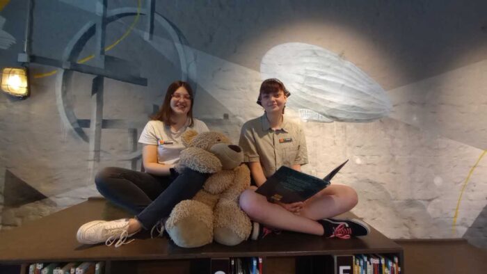 Unsere Beiden FaMI Auszubildenden Marie Bonfig und Rosalie Schöpf sitzten in der Stadtteilbücherei Hubland und lesen gemeinsam mit Hubärt, einem großen Teddybär ein Buch.