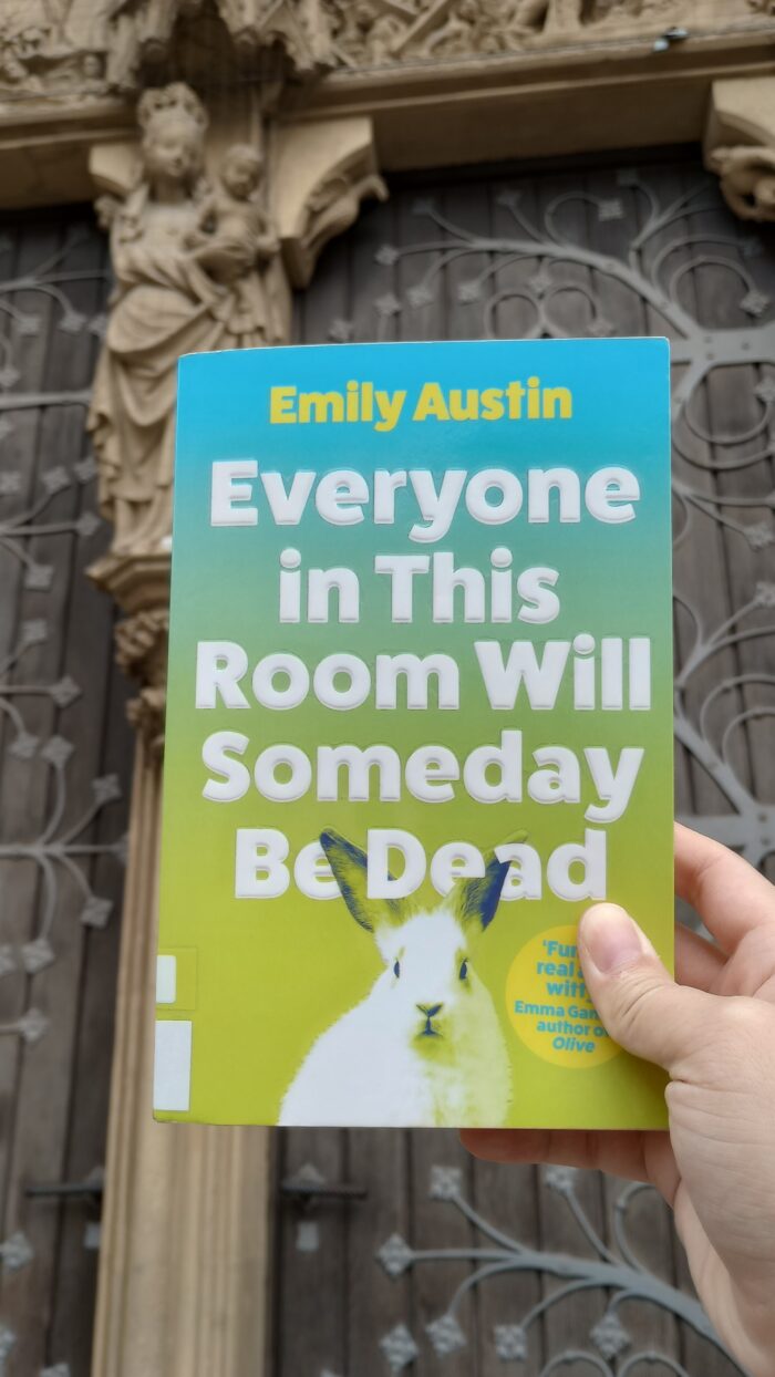 Das Buch "Everyone in this room will someday be dead" von Emily Austin wird von einer Hand vor eine Kirchentür gehalten.