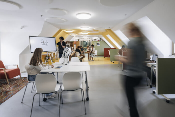 Eine Aufnahme aus dem "Level3", einem Raum für Digitalisierung. Links stehen zwei Tische, an denen eine Person sitzt und zu einer Person sieht, die etwas auf ein Whiteboard schreibt. Rechts läuft eine Person vorbei. Im Hintergund sind Menschen zu sehen, die sich mit den verfügbaren Geräten beschäftigen.