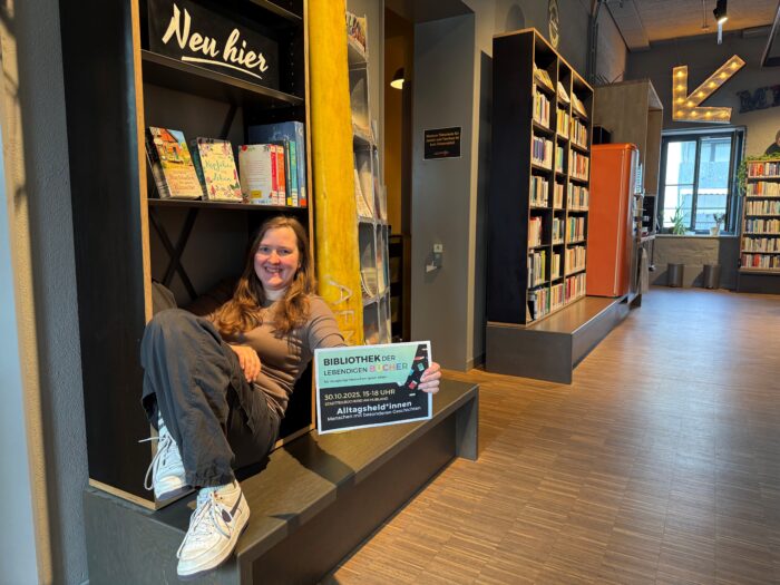 Eine junge Frau sitzt in einem Regal in der Stadtteilbücherei Hubland und hält ein Veranstaltungsplakat für die "Bibliothek der lebenden Bücher" am 30. Oktober 2025.