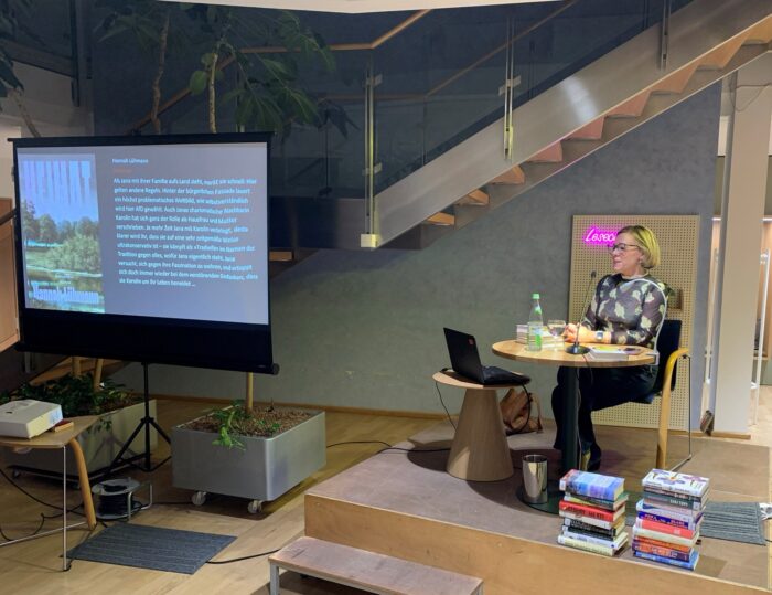 Referentin Isabel Fraas stellt im Lesecafé die Bücher der Saison vor. SIe sitzt rechts an einem Tisch, links läuft eine Präsentation über den Beamer.