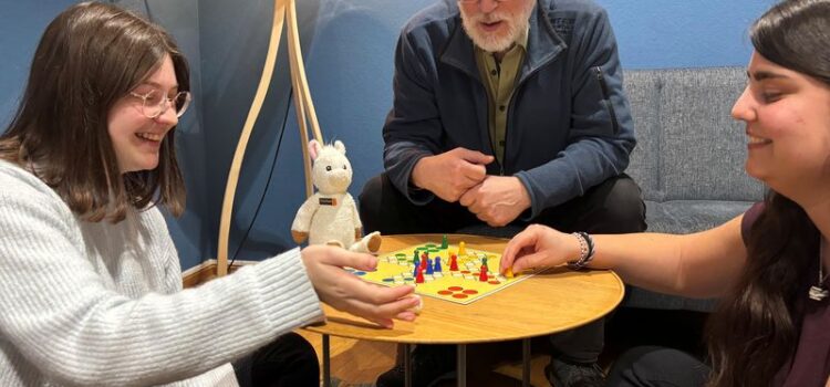 Auf dem Bild befinden sich zwei junge Frauen, die mit einem älteren Mann und einem Kuscheltierlama um einen Tisch sitzten und "Mensch ärgere dich nicht" spielen