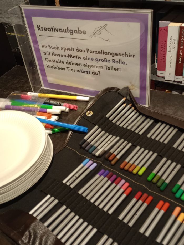 Zu sehen ist eine dunkelbraune Oberfläche auf der ein Schild mit folgender Aufschrift steht: "Kreativaufgabe: Im Buch spielt das Porzellangeschirr mit Hasen-Motiv eine große Rolle. Gestalte deinen eigenen Teller: Welches Tier wärst du?" Daneben sind ein Stapel weißer Pappteller, FIlzstifte und Buntstifte.