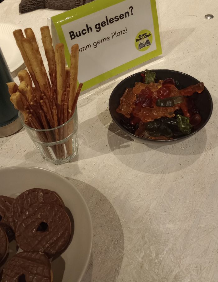 Zu sehen ist ein weißer Holztisch, auf dem Schälchen mit Keksen, Salzstangen und Gummibärchen stehen, sowie ein Schild mit der Aufschrit "Buch gelesen? Nimm gerne Platz!".