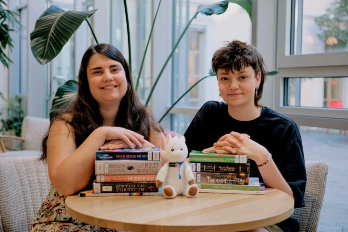 Zu sehen sind zwei Mitarbeiterinnen der Stadtbücherei mit zwei Stapeln queerer Bücher.