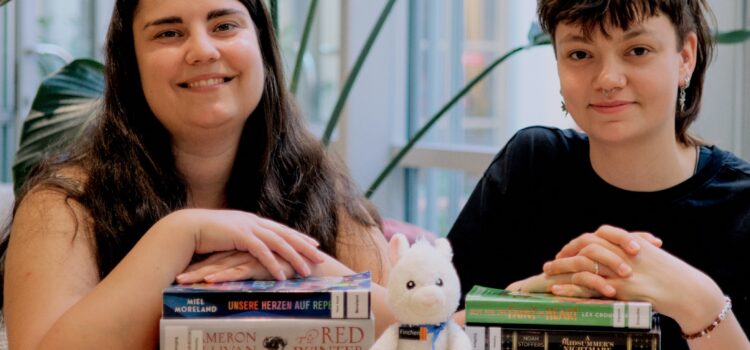 Zu sehen sind zwei Mitarbeiterinnen der Stadtbücherei Würzburg mit zwei Bücherstapeln. Daneben sitzt ein Plüsch-Lama.