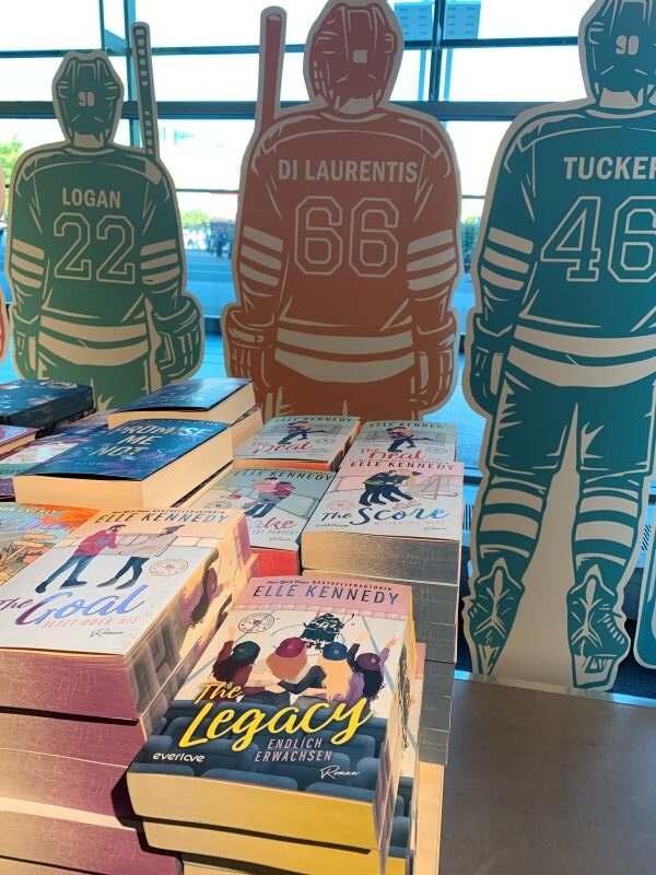 Die Romance-Bücher der Off-Campus -Reihe von Elle Kennedy liegen auf einem Tisch. Dahinter befinden sich Pappaufsteller mit den Eishockey-Spielern der einzelnen Bücher.